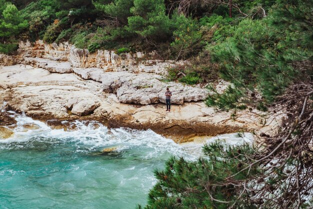 Mann steht mit Telefon am felsigen Strand und blickt auf stürmische See