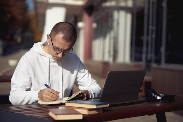 Mann studiert am Laptop und schreibt in ein Notizbuch, während er auf der Straße an einem Tisch sitzt. Soziale Distanzierung während des Coronavirus