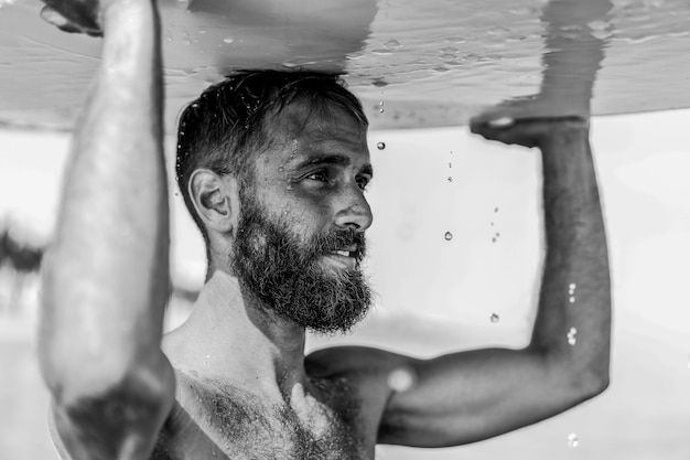 Foto mann-surfer, der sein surfbrett bei sonnenaufgang trägt hipster-männchen in hemdlosem stehen am strand lifestyle- und extremsportkonzept konzentrieren sie sich auf die augen schwarz-weiß-bearbeitung