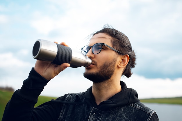 Mann trinkt Wasser aus wiederverwendbarer Thermoflasche aus Stahl auf dem Hintergrund eines verschwommenen bewölkten Himmels