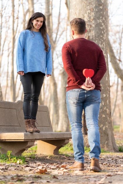 Mann überrascht seine Freundin mit einem herzförmigen Geschenk Sie sitzt auf einer Bank im Park