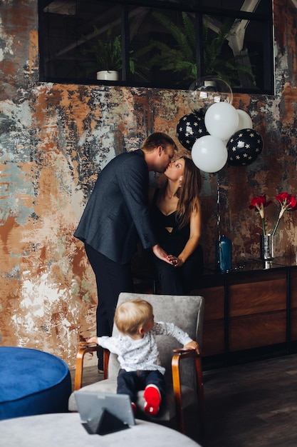 Mann und Frau, die zusammen stehen, küssen und Händchen halten Familie feiert mit Luftballons und Blumen und posiert im Wohnzimmer Kleines Kind, das auf dem Stuhl sitzt und auf seine Eltern zurückblickt