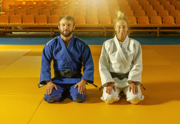 Mann und Frau im blau-weißen Kimono mit schwarzem Gürtel sitzen auf dem Boden und meditieren in der Sporthalle. Orientalische Kampfkunst, Judo