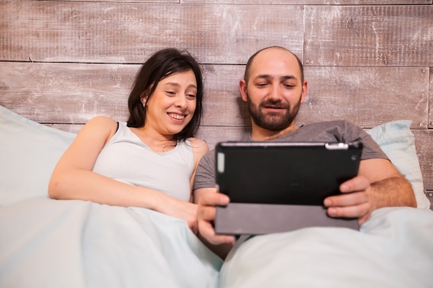 Mann und Frau im Pyjama lachen beim Ansehen eines lustigen Videos auf dem Tablet-Computer.