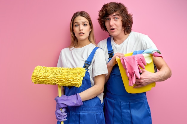 Mann und Frau in Overalls und Gummihandschuhen betrachten Kamera halten Reinigungswerkzeuge, die über rosa Studiohintergrund isoliert werden