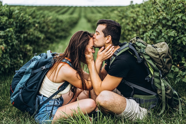 Mann und Frau mit Rucksäcken auf dem Rücken sitzen umarmend auf dem grünen Gras zwischen den Johannisbeersträuchern. Wandern auf den Johannisbeerplantagen auf dem Land