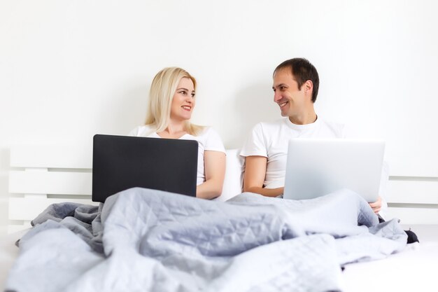 Mann und Frau sitzen morgens auf dem Bett und haben Spaß, lachen, arbeiten an Laptop-Onile-Ausbildung, Online-Freiberufler-Job, lächeln glücklich, Familie zusammenleben, Schlafzimmer, Pyjama tragen