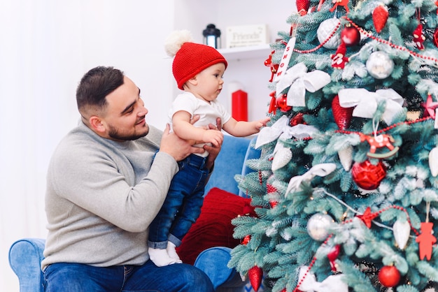 Mann und süßes Baby sind bereit, neues Jahr zu feiern. Glücklicher Vater spielt mit seinem kleinen Sohn nahe Weihnachtsbaum am Weihnachtsabend.