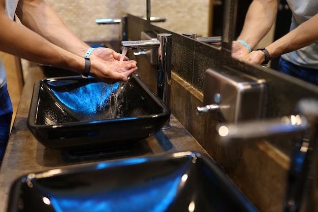 Mann waschen seine Hand an einem öffentlichen Ort mit Wasserhahn mit Wasser. Die Toilette hat ein modisches schwarzes Waschbecken und einen Metallhahn.