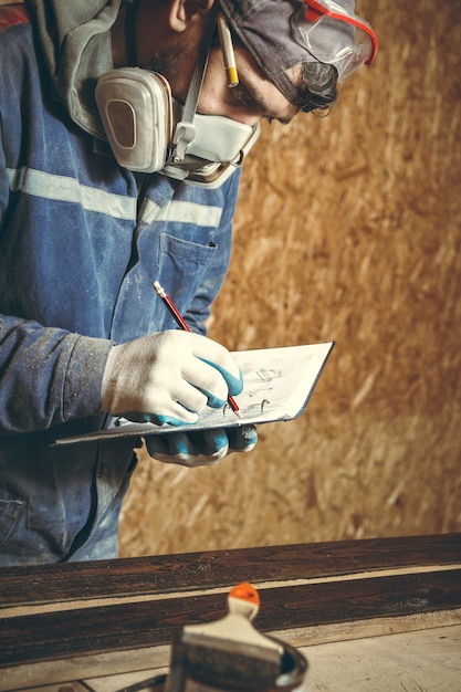 Mann Zimmermann in seinem Heimstudio arbeitet mit Holz und zeichnet Skizzen mit Bleistift in ein Notizbuch