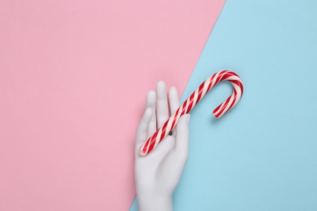 Mannequinhand, die weihnachtliche gestreifte Zuckerstange auf einem blaurosa Hintergrund hält