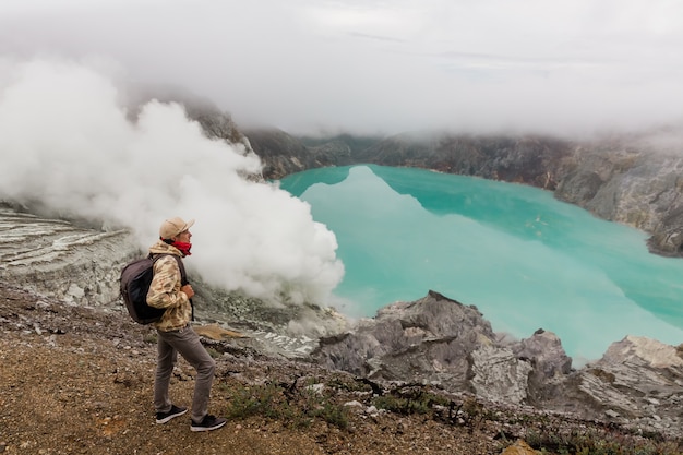 Manntourist betrachtet den Schwefelsee auf dem Ijen-Vulkan auf der Insel Java in Indonesien. Wanderermann mit Rucksackreise auf Spitzenberg, Reisekonzept