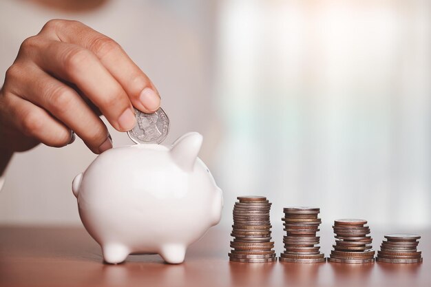 Foto la mano ahorrando una moneda en la alcancía con un paso de monedas y dinero en la mesa de madera