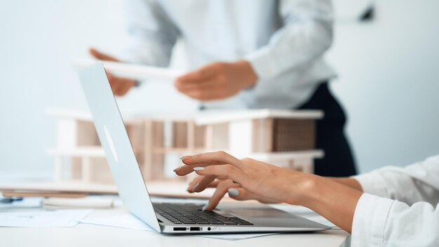 La mano del arquitecto usando la computadora portátil mientras su compañero de trabajo revisa el modelo de casa Inmaculada