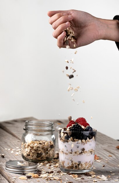 Mano arrojando cereales sobre un vaso con yogur y frutas del bosque. concepto de desayuno saludable