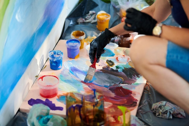 La mano del artista con guantes negros sostiene el pincel y elige el color de la paleta de colores en el festival de arte