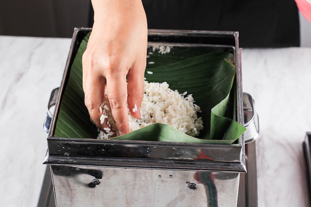 Mano asiática femenina poner arroz pegajoso en la vaporera de acero inoxidable con hoja de plátano, proceso de preparación de lemper, postre asiático o refrigerio