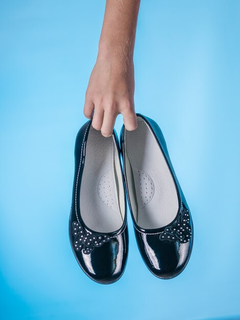 La mano del bebé sostiene elegantes zapatos de cuero sobre fondo azul. Zapatos de mujer de cuero elegantes y de moda.