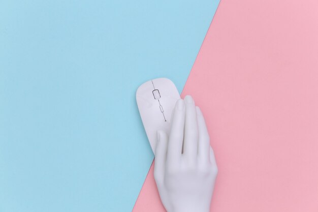 La mano blanca del maniquí que sostiene el mouse de la PC sobre fondo rosa pastel azul. Vista superior