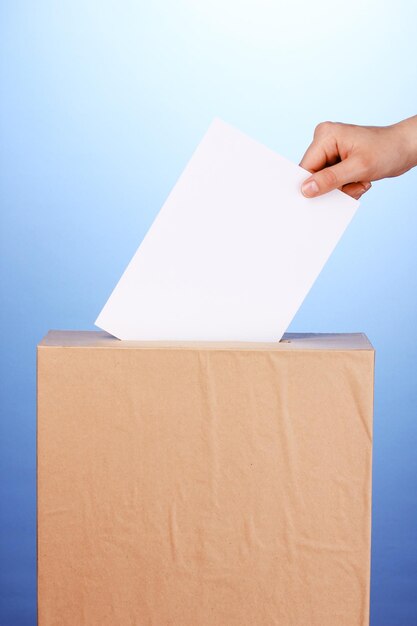 Mano con boleta de votación y caja sobre fondo azul.