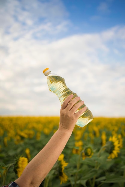 Una mano con una botella de aceite de girasol dorado levantada contra el fondo de un campo de girasoles en flor en un soleado espacio de copia