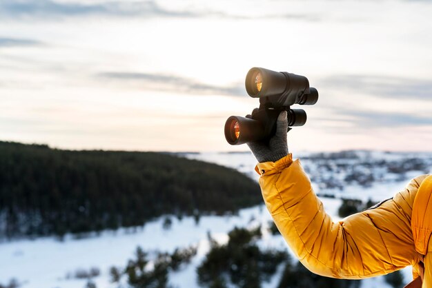 mano en chaqueta amarilla sosteniendo binoculares contra la vista del bosque invernal río nevado Ecología de observación de aves