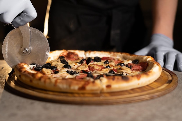 La mano del chef con un cuchillo corta pizza caliente con aceitunas y salami en la mesa sobre una tabla de madera en una pizzería. Productos horneados listos para la entrega.