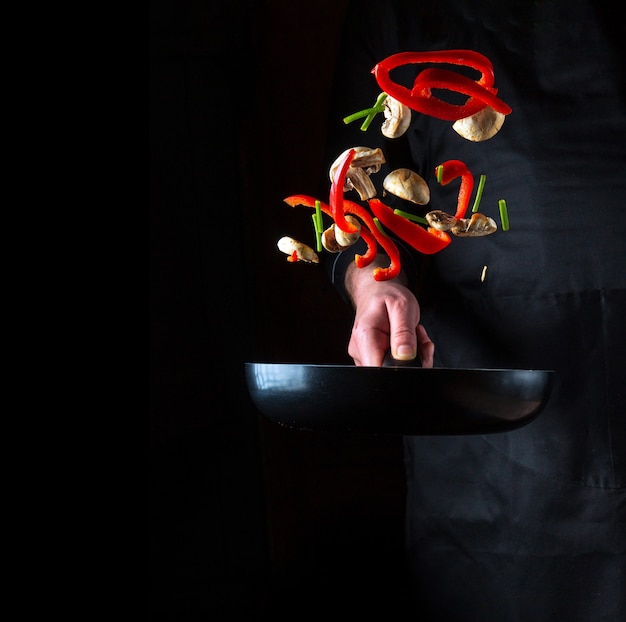 La mano de un chef profesional arroja trozos de verduras y setas en una sartén sobre un fondo negro. Concepto de cocina de restaurante o cafetería. Espacio publicitario gratuito