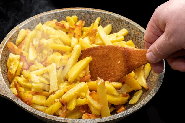 Foto mano del chef revolviendo con una espátula de madera deliciosos trozos de patata frita caliente dorada crujiente en una vieja sartén de metal de cerca. cocinar patatas fritas caseras en una sartén