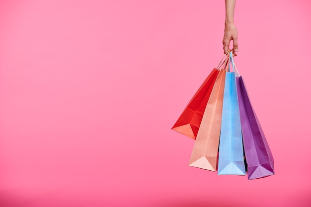 Mano del comprador sosteniendo un montón de coloridas bolsas de papel o llevándolas después de comprar sobre fondo rosa