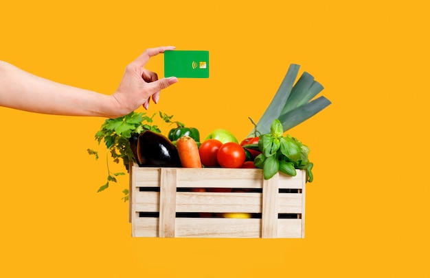 Mano de compras de alimentos orgánicos con tarjeta de crédito por encima de la caja con verduras frescas