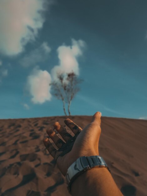 Foto mano cortada de un hombre gestando contra un árbol en una duna de arena contra el cielo