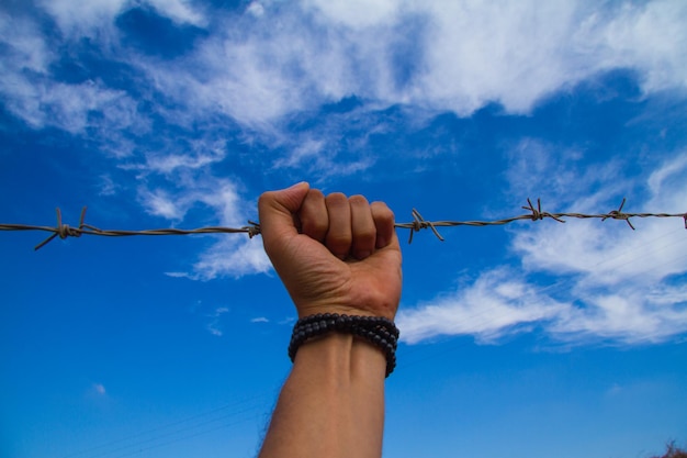 Mano cortada de un hombre sosteniendo alambre de púas contra el cielo azul