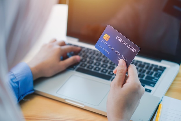 Mano cortada de una mujer de negocios sosteniendo una tarjeta de crédito mientras usa una computadora portátil en la oficina