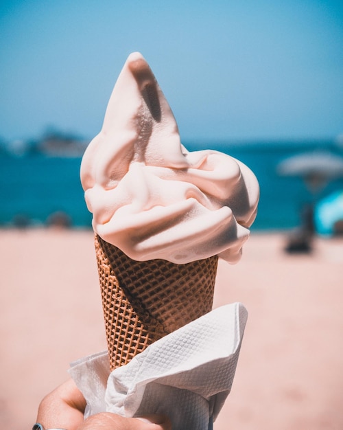 Mano cortada de una mujer sosteniendo un cono de helado en la playa contra un cielo azul claro
