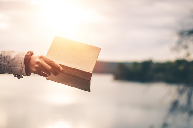 Foto mano cortada de una mujer sosteniendo un libro junto al lago contra el cielo durante la puesta de sol