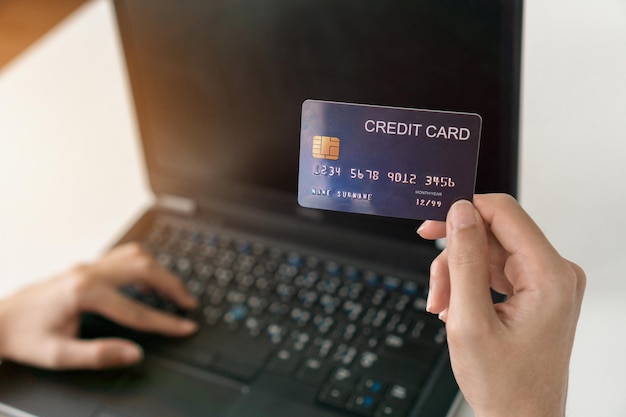 Mano cortada de una persona que sostiene una tarjeta de crédito mientras usa una computadora portátil para comprar en línea
