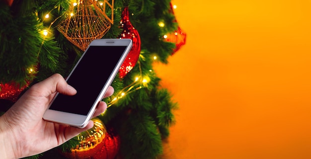 Mano cortada de una persona usando un teléfono móvil por un árbol de Navidad iluminado