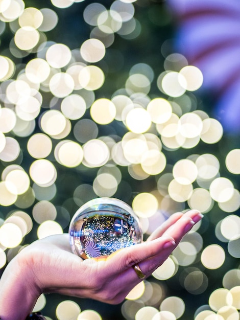 Foto mano cortada sosteniendo una bola de cristal contra luces iluminadas durante la navidad por la noche