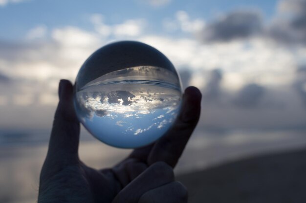 Mano cortada sosteniendo una bola de cristal en la playa contra el cielo