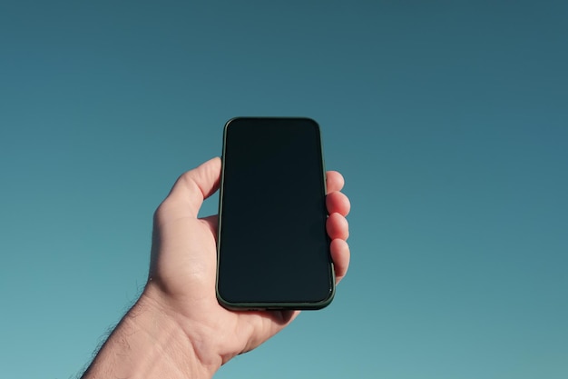 Foto mano cortada sosteniendo un teléfono inteligente contra un cielo azul despejado