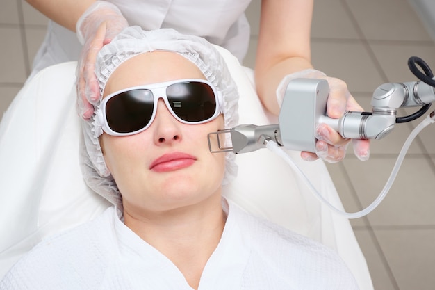 La mano de la cosmetóloga sostiene el láser de neodimio en la cara del paciente con gafas haciendo el enfoque selectivo del procedimiento cosmetológico