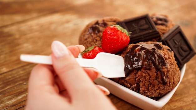 Mano con una cuchara. Helado de chocolate casero con chocolate y fresas. Cuenco blanco y fondo de madera