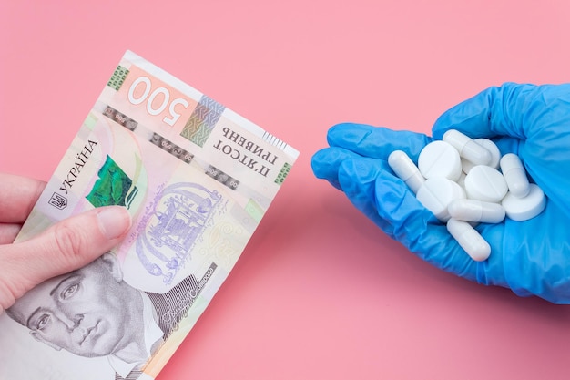 La mano da un billete de quinientos hryvnia hacia una mano en un guante médico con una pastilla sobre un fondo rosa. El concepto de comprar medicamentos caros.