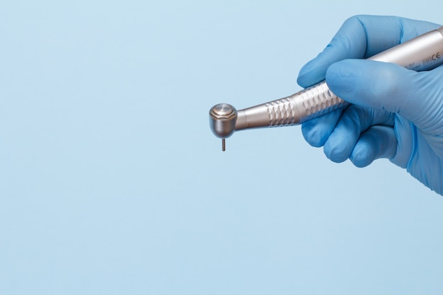 La mano del dentista en un guante de látex con pieza de mano dental de alta velocidad sobre fondo azul. Concepto de herramientas médicas.