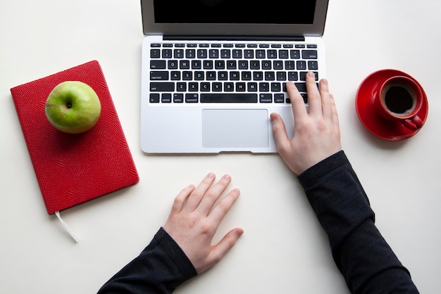 Mano derecha del hombre en el teclado de la computadora portátil y mano izquierda en la mesa blanca con una taza de café roja y un cuaderno, manzana verde