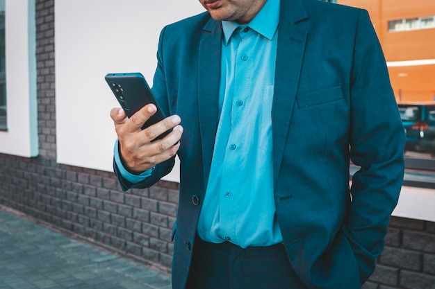 Mano ejecutiva usando un teléfono inteligente en la calle con un edificio de oficinas en el fondo Un hombre de negocios con traje azul mira un teléfono inteligente en su mano Trabajo de negocios sobre la marcha