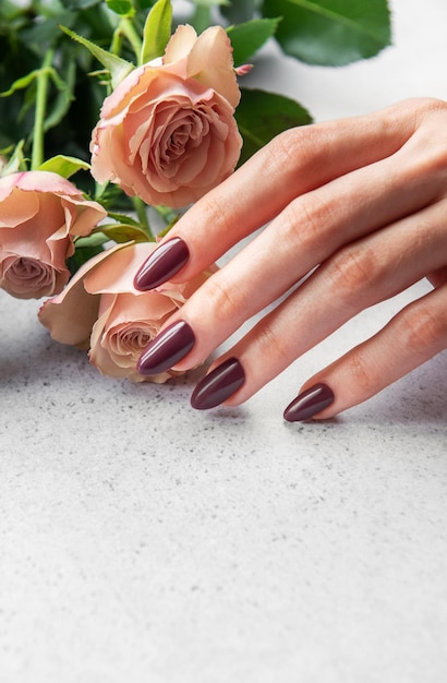 Mano elegante con manicura oscura descansando al lado de delicadas rosas rosadas en una superficie clara