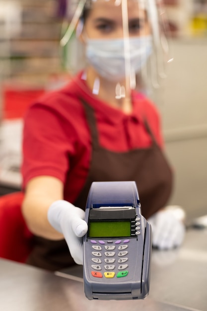 Mano enguantada de cajera en uniforme, pantalla protectora y máscara con terminal de pago en el mostrador de pago en el supermercado