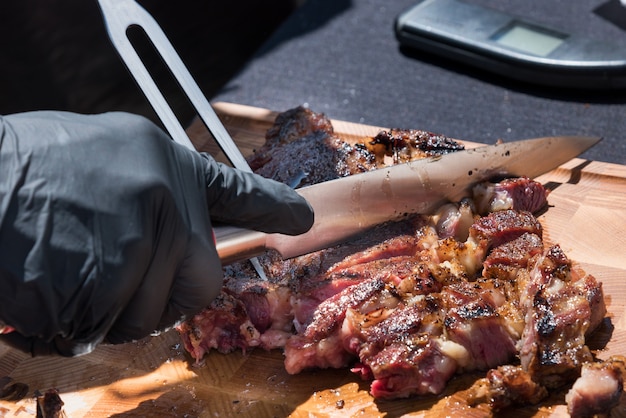 Mano enguantada de un chef tallando una porción de carne de costilla a la parrilla de la barbacoa en una tabla de cortar de madera en un primer plano de la comida